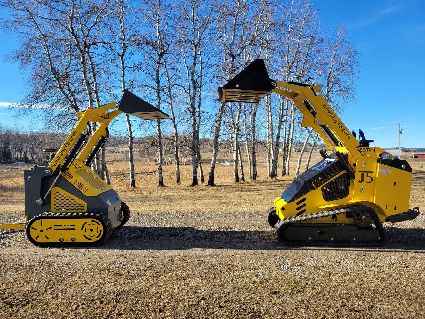 Side-by-side comparison of JoyT5 and JoyT5 SSTM4 mini skid steer loaders with raised buckets, displayed outdoors on a clear day.