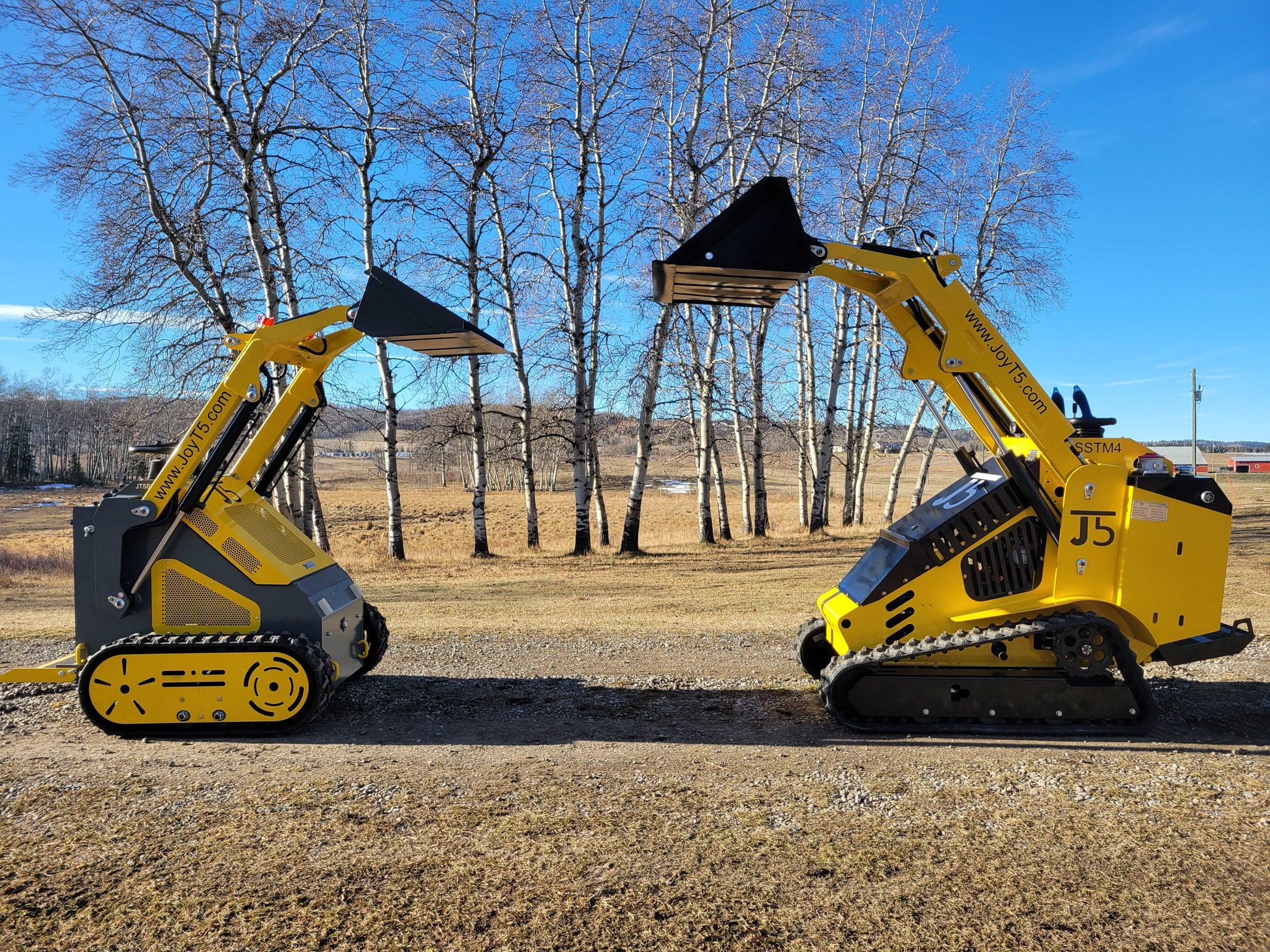 Side-by-side comparison of JoyT5 and JoyT5 SSTM4 mini skid steer loaders with raised buckets, displayed outdoors on a clear day.