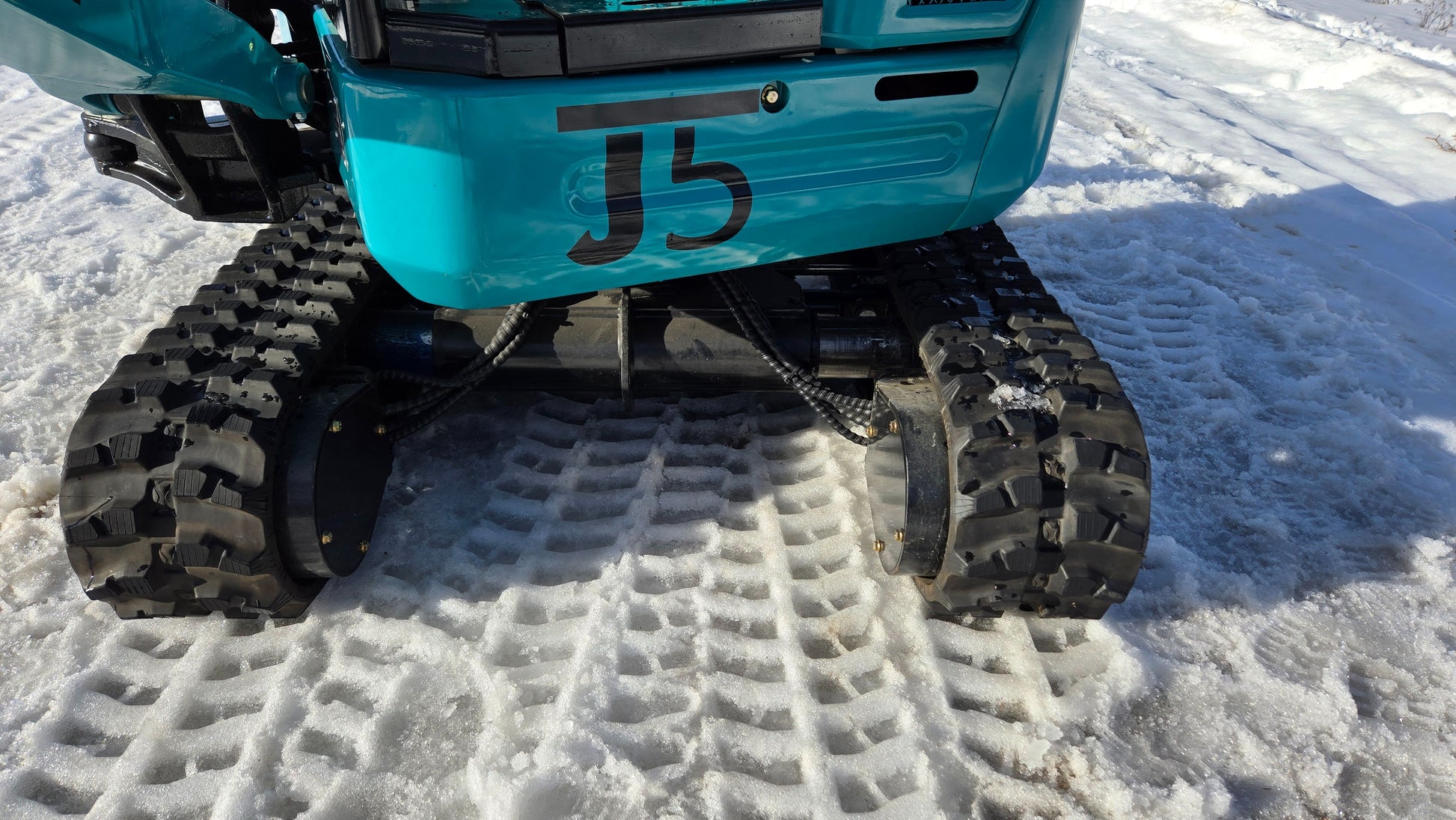 Rear undercarriage view of JoyT5 JT5200 mini excavator showing dual rubber tracks, hydraulic motors, and frame on snow.
