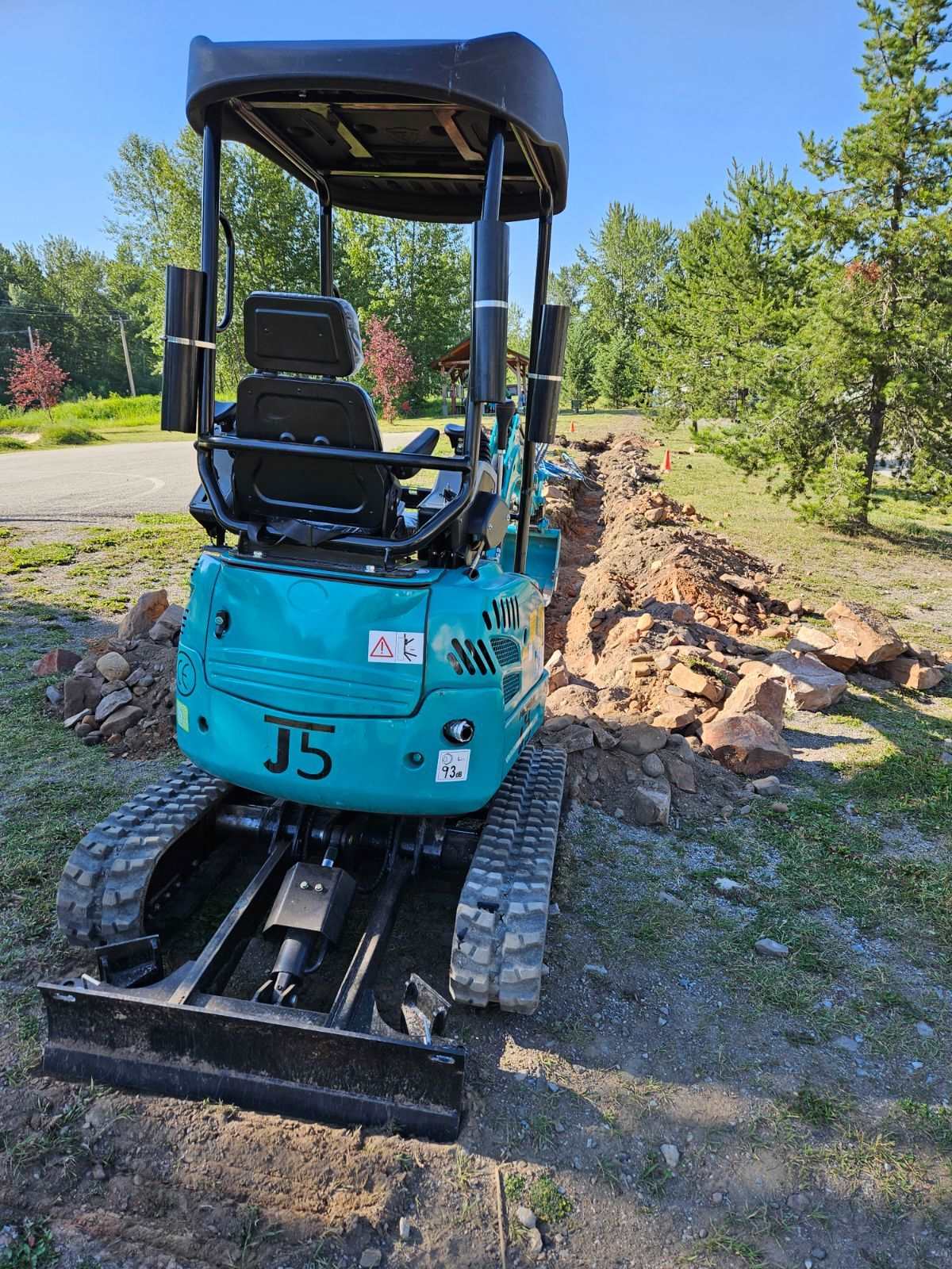 Rear view of JoyT5 JT5200 2 ton mini excavator with Kubota engine digging trench and moving soil with dozer blade. Compact hydraulic excavator designed for landscaping, utility, and construction projects.
