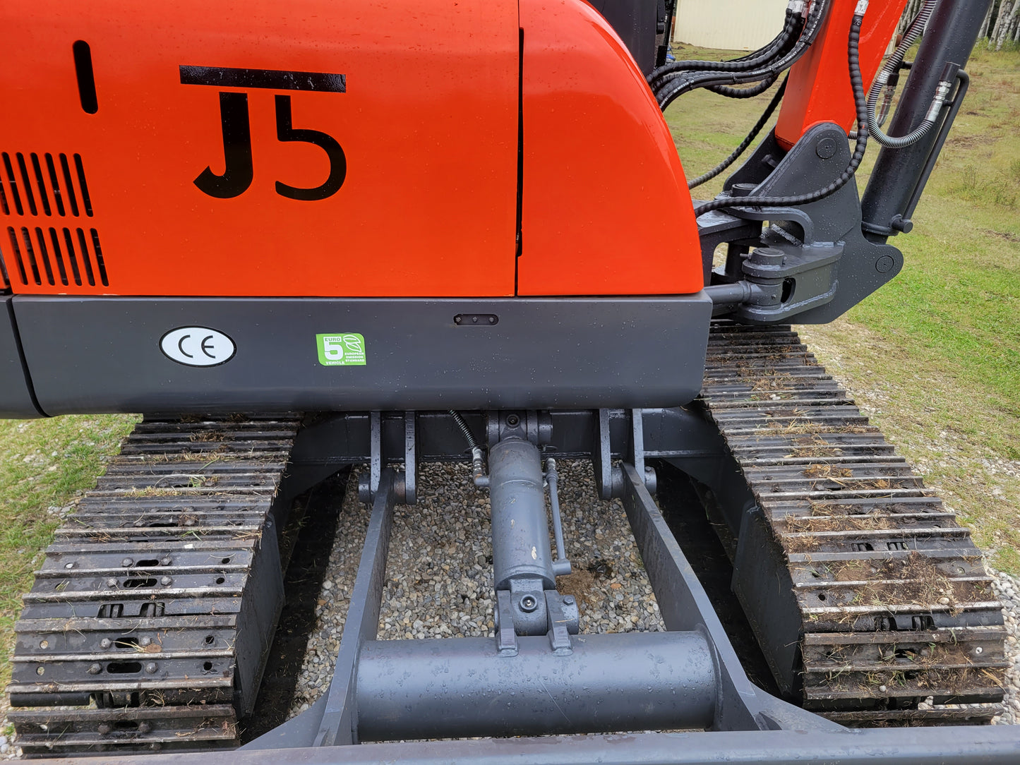 Close-up view of JT5600 mini excavator undercarriage showing hydraulic cylinder, steel tracks, and part of body panel with CE certification and eco-friendly label