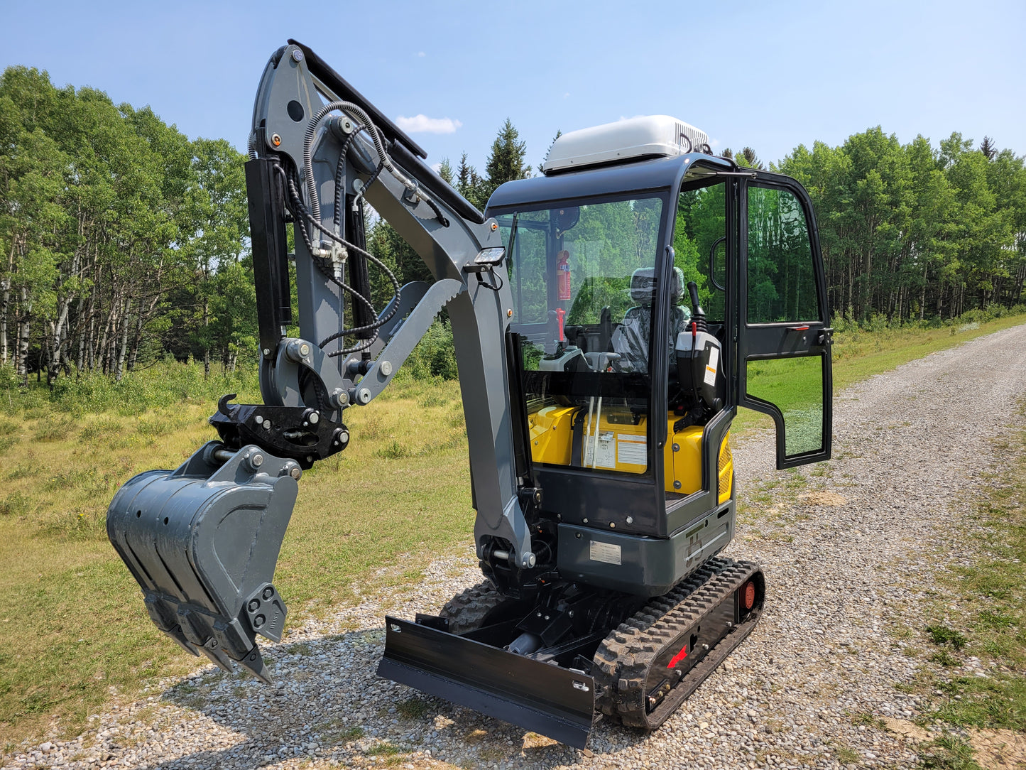 Compact mini excavator with cabin and digging bucket, parked outdoors on gravel road near green forest, showing detailed structure and design.