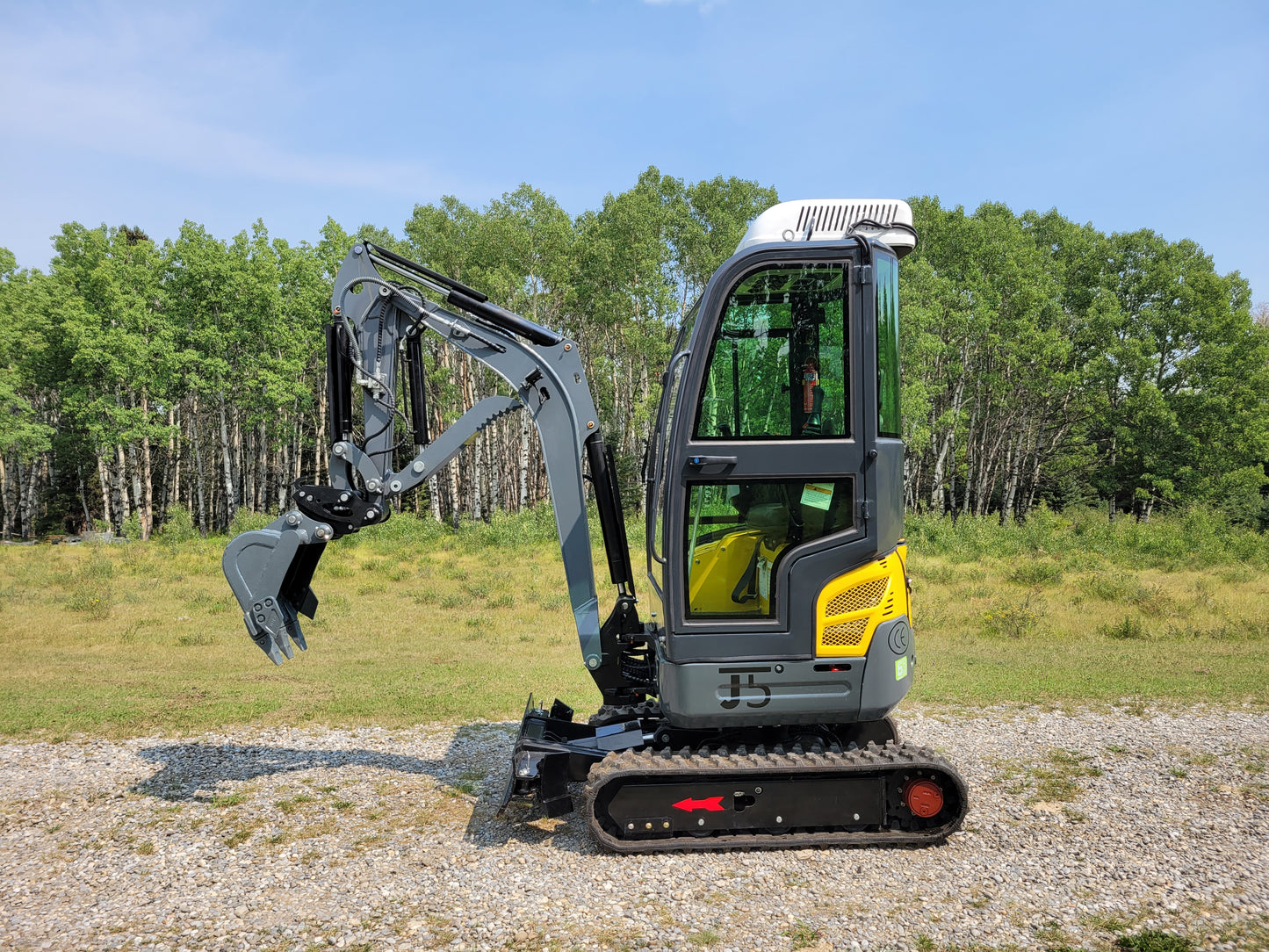 Side view of compact mini excavator with enclosed cabin and digging bucket, parked on gravel near green forest background.