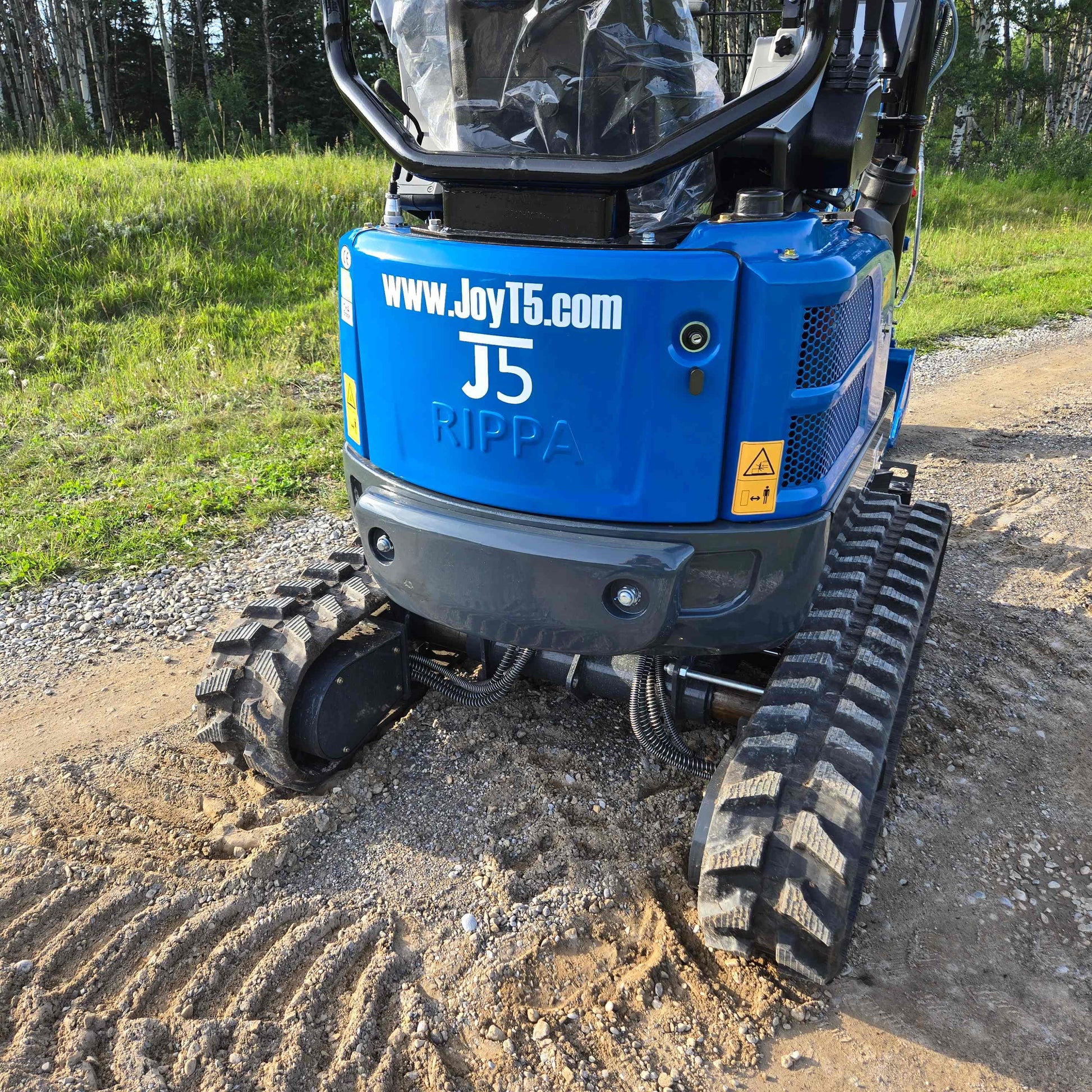 Rear view of JoyT5 Rippa R18 Mini Excavator for sale with branding and rubber tracks visible, positioned on a gravel and dirt road surrounded by greenery.