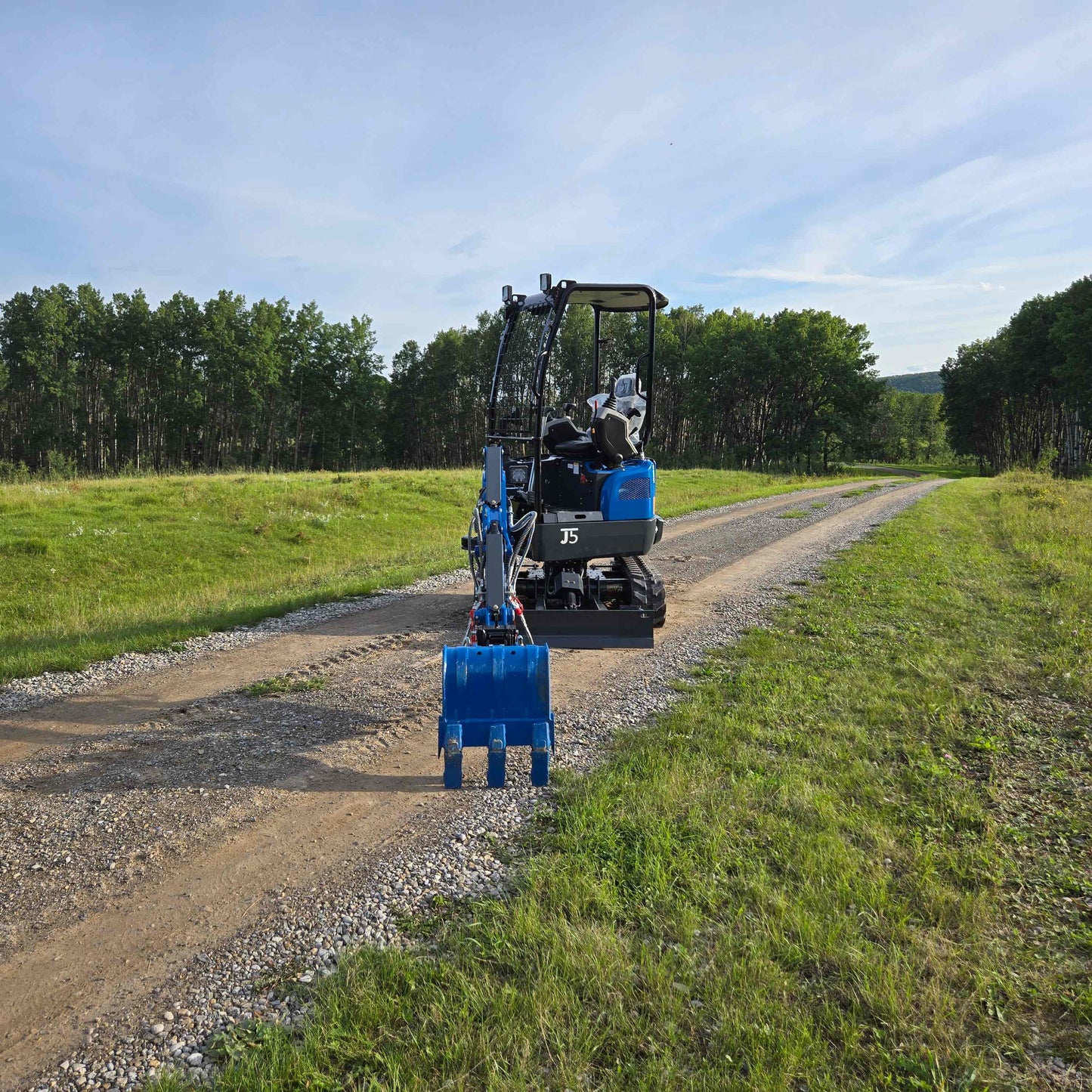 Rear view of JoyT5 Rippa R18 Mini Excavator with blue digging bucket on a gravel road surrounded by greenery