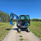 Front view of the Rippa R22 Mini Excavator for sale with cabin door open and digging arm extended, parked on a gravel path surrounded by grass and trees.
