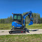 Full side view of Rippa R22 Mini Excavator for sale on gravel road with bucket attachment raised, in front of trees.
