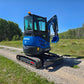 Rear side view of the Rippa R22 Mini Excavator for sale with front blade and digging arm extended, parked on a gravel path with trees in the background.
