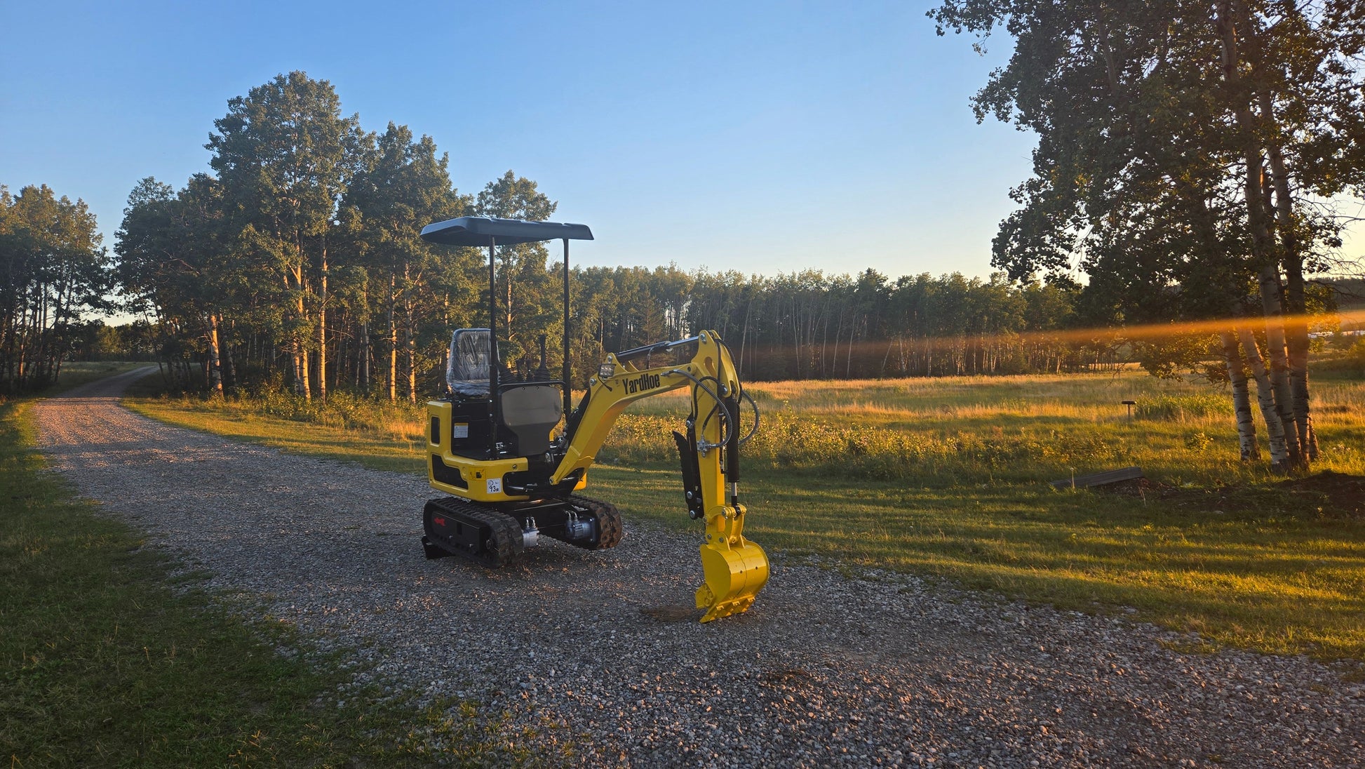 YardHoe YH12 mini excavator parked on a gravel path with digging bucket lowered, trees and sunset in the background.
