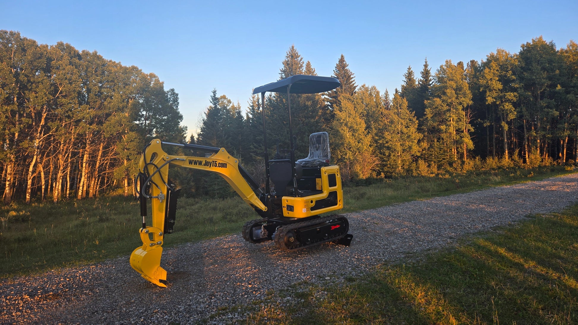 Yellow YardHoe YH12 mini excavator with canopy parked on a gravel road, surrounded by green forest trees at sunset.
