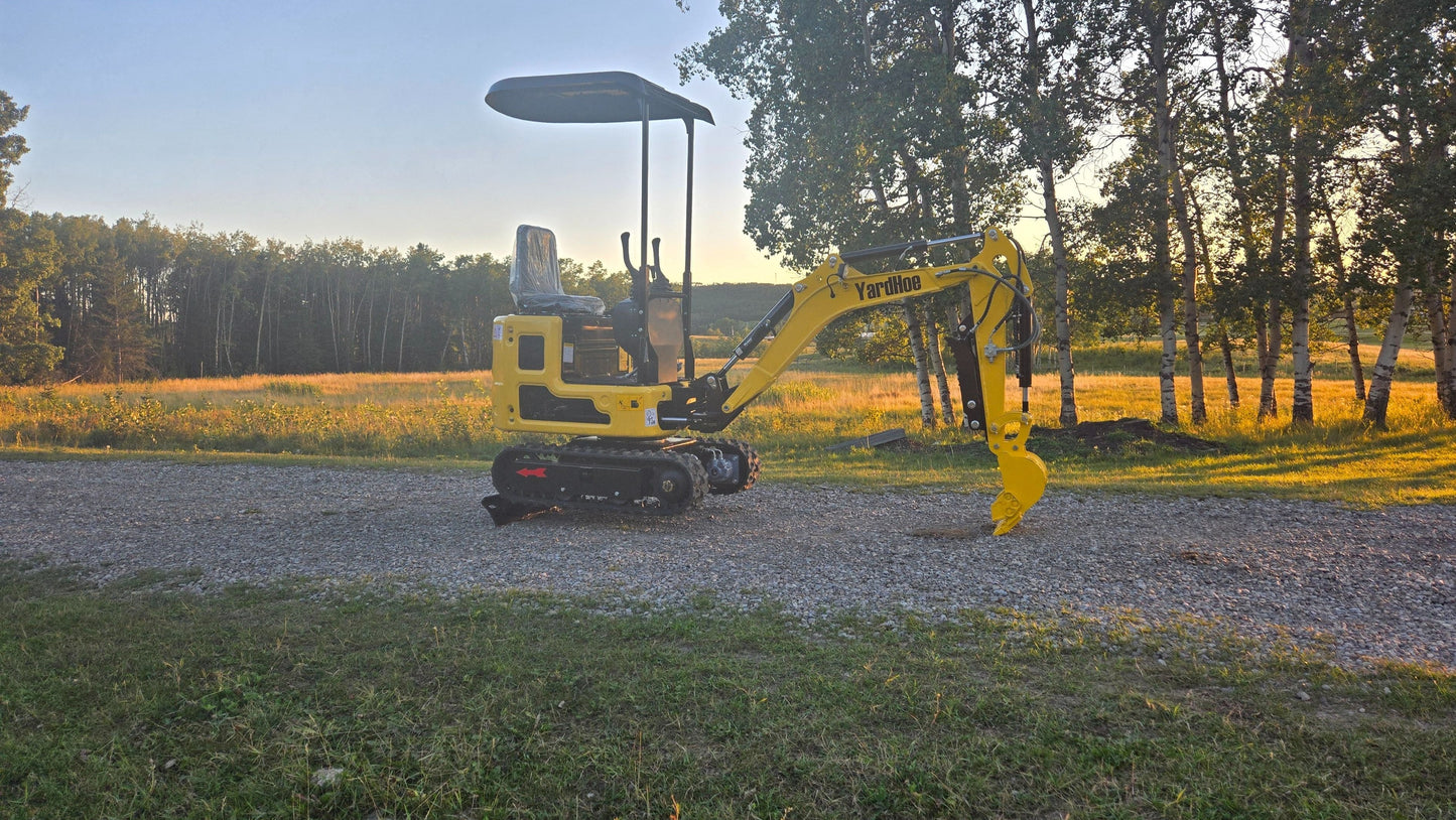 YardHoe YH12 mini excavator parked on a gravel road with digging arm lowered, in front of trees at sunset.
