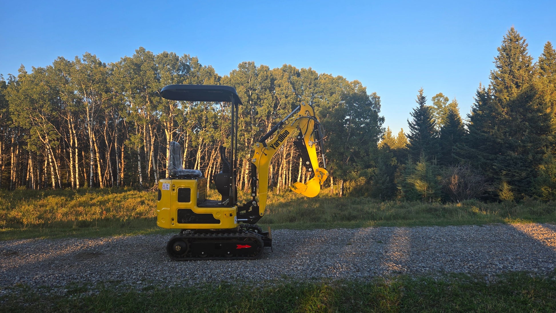 Side profile of YardHoe YH12 mini excavator on gravel road with forest trees in the background.
