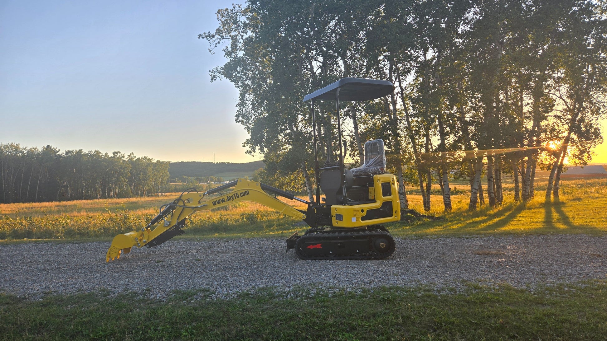 Side view of YardHoe YH12 mini excavator parked on gravel road with sunset and trees in the background.

