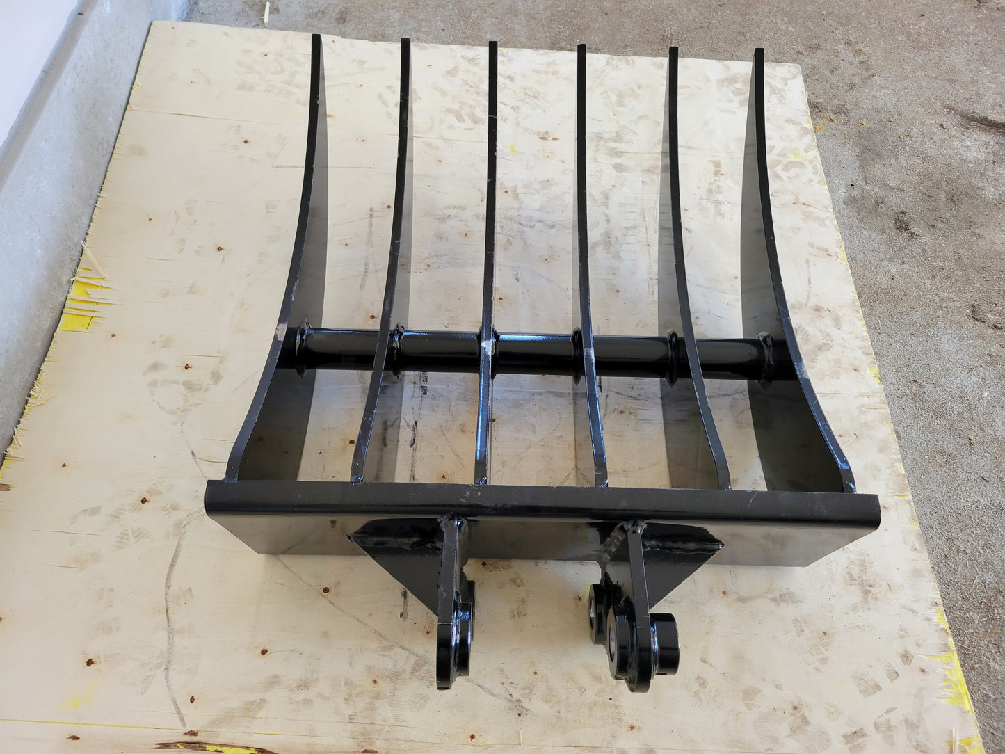 Mini excavator rake attachment front view showing multiple curved steel tines for landscaping site cleanup and heavy construction work in Canada